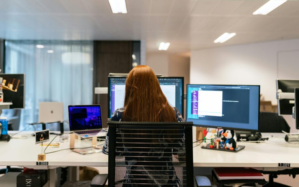 Back view of a female software engineer working at a multi-monitor setup in an office.